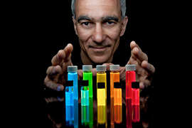 Moungi Bawendi's head and hands appear against a black background and over a rainbow-colored row of vials
