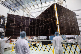 Workers in white protective clothing watch as the solar arrays for Psyche spacecraft are retracted. The combined solar arrays are about the size of a tennis court.