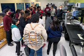 While about 30 students look on, an instructor points to a machine in a lab filled with oscilloscopes and other equipment.