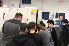 An instructor shows students a “Robocut” machine in factory, as 7 students look on.