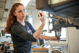 Liberty Ladd wears safety goggles while operating a large drill machine in a workshop.