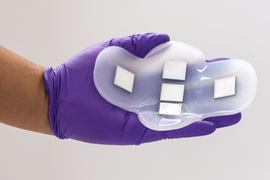 A gloved hand holds a soft, flexible patch made of silicone. It has 5 square sensors positioned in a cross.
