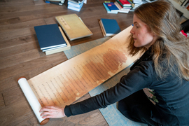 Wiebke Denecke sits on the floor amongst books and examines a long unfurled scroll with Chinese writing.