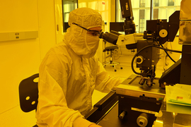 Peter Satterthwaite looks into a microscope while wearing a full-body suit in the yellow-lit MIT.nano lab.