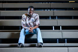 Kwesi Afrifa sits on the bleachers of a theatre with hands clasped. The rows of bleachers create strong horizontal lines.