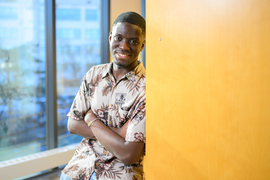 Kwesi Afrifa smiles while leaning against a yellow wall, with windows in background.