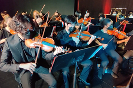 Three violinists are in foreground, wearing masks and playing as part of a larger ensemble. About 15 other musicians are seen in the background.