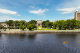 Drone photo shows the Charles River and MIT campus, with Killian Court in the middle, on a sunny day. Two people are in a kayak.
