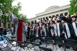 A professor in academic regalia stands on a chair and takes a group selfie with about 30 excited graduates wearing caps and gowns.