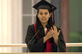 Penny Brant, wearing cap and gown, speaks at a podium and holds up her hand with her graduation ring.