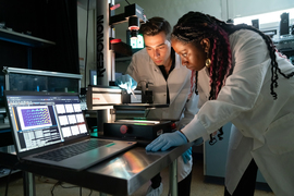 Eunice Aissi and Alexander Siemenn look into at a back-lit microscope in laboratory.