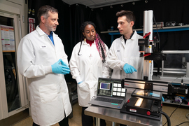Tonio Buonassisi, Eunice Aissi, and Alexander Siemenn stand near laptop and lit-microscope in laboratory.