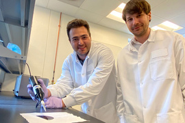 Two men in a lab in white suits gesturing at a large wafer on the table.