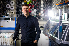 Skylar Tibbits stands in the lab with a wall of colorful spools behind him.