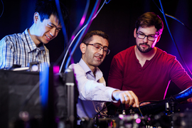 Three people observe a table of equipment, with pink and blue lighting.