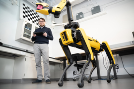 Luca Carlone holds a controller in background, with a yellow dog-type robot in foreground.