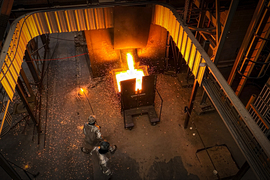 A blast of molten steel causes dazzling sparks as two people in protective suits, one with a long pole, stand back.