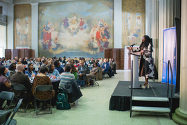 Oluwadara Deru speaks at a podium in a large hall, while audience members listen at tables