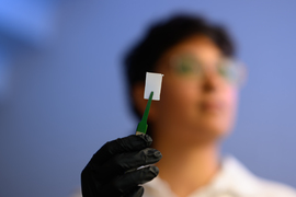 With black gloves Palak Patel holds up a white rectangle the size of a postage stamp, gripped with tweezers. Patel's blurred face is in the background.