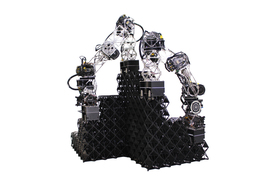 Two robot arms stand on top of a voxel structure as they continue to build it up.