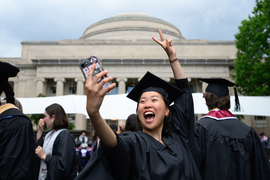 A female student in cap and gown takes a selfie in front of the Great Dome