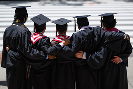 Five young men in caps and gowns stand side by side, facing away from the camera, with arms around each other.