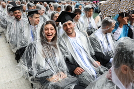 Rows of seated graduates wear clear ponchos and smile despite rain