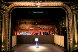 The stage design features a golden arch opening on a giant stage with bare wooden walls. A single dancer performs in a spotlight. On top of the walls are musicians against a rough rocky background.