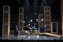 Two men in formal wear sing while one throws trash and paper in the air. The stage is mainly constructed with large rectangular boxes that have green typography projected onto them.