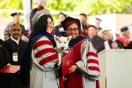 Michelle Quien holds her dog, a white terrier, while both are dressed in graduation regalia and face Paula Hammond.