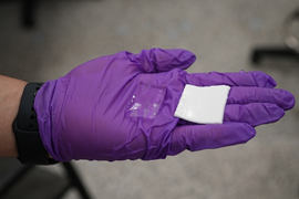 A gloved hand holds two square examples of hydrogel, one clear and one white.