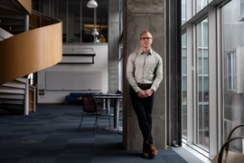 Brian Hedden stands in a lounge with large glass walls at right and a wooden spiral staircase at left
