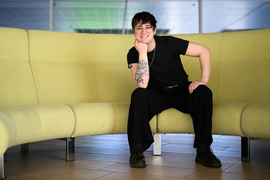 Shannon Knight portrait, seated on curved yellow couch. An intricate forearm tattoo with leaves and faces is visible.