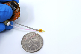 A small circuit board and pump connected to a slender gold-colored device beside a coin.