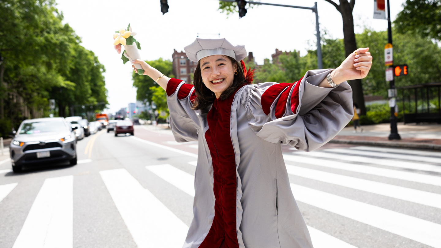 An MIT graduate in PhD graduation regalia smiles brightly as she crosses the street and holds a bouquet of flowers.