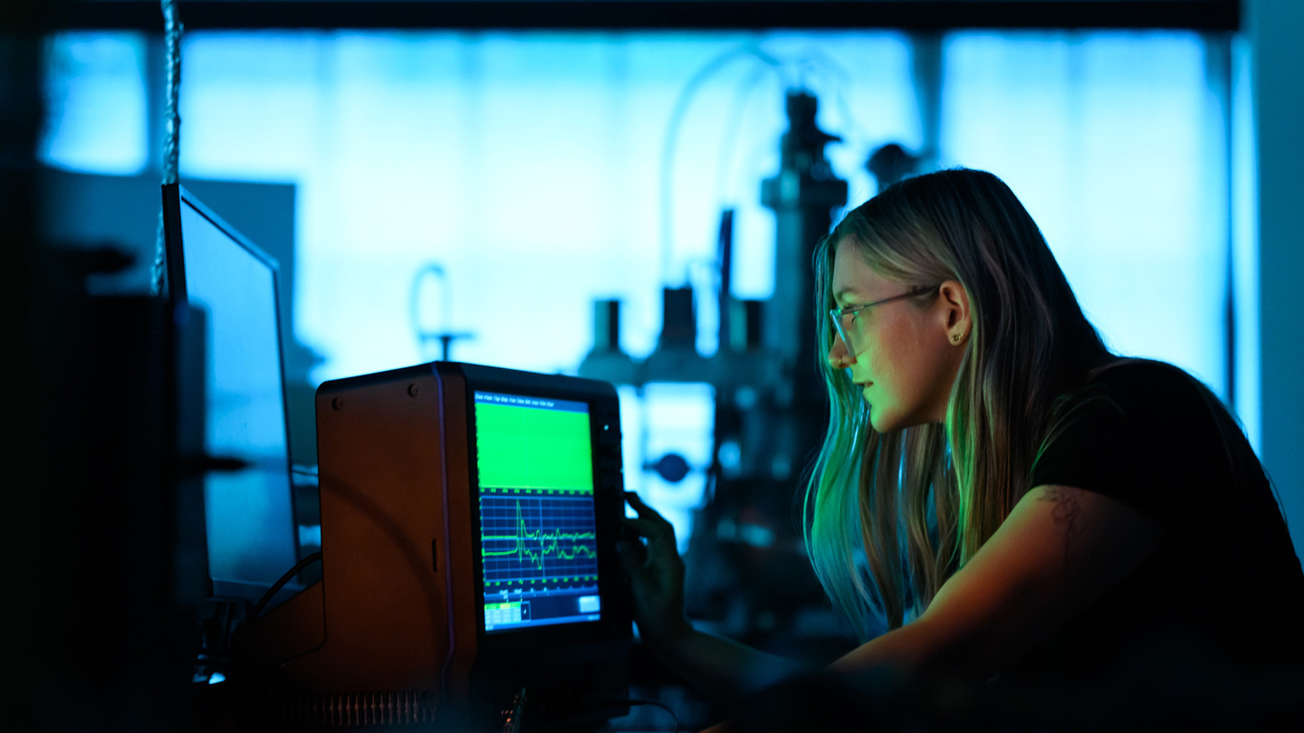 Lanie McKinney looks at glowing green waveforms on an oscilloscope in the lab.