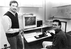 Mandayam Srinivasan, director of MIT's Touch Lab, stands next to a monitor displaying a virtual rendition of a liver. Research Scientist Cagatay Basdogan is manipulating the liver with a robotic device that translates the touch and feel of virtual objects to the user. The work could lead to surgical training based on virtual reality.
