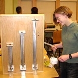 MIT freshman Ellen Cappo demonstrates a column buckling exhibit. By loading the white bucket on the right the same load is transmitted to each of the three columns. The exhibit shows the relationship between column weight and height, and explains why a tree can only grow so tall. Professor Lorna Gibson taught a freshman seminar in which students put together this and other interactive exhibits on ...