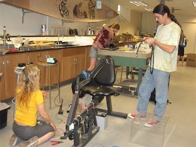 In the MIT Course 1.102 laboratory, lecturer Jessica Banks (kneeling) and students Kendra Johnson '09 and Sebastian Figari '09 get ready to secure the laptop to the swivel arm of the exercise bike.