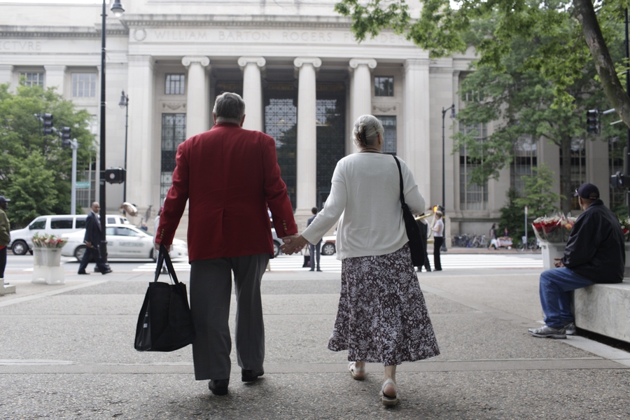 An MIT alumnus &#8212; whose red coat signifies he graduated 50 years or more ago &#8212; and his wife walk toward MIT&#39;s main entrance at 77 Massachusetts Avenue.
