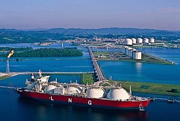 Tanker with several large domes floats in a bay where large gas tanks also sit