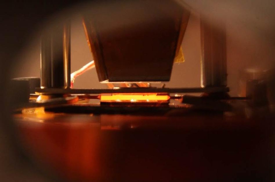 Optical image of the vacuum-enclosed device illustrating the energy conversion processes in a nanophotonic solar thermophotovoltaic device: Sunlight is converted to useful thermal emission, and ultimately electrical power, via a hot absorber-emitter (at center, glowing orange).