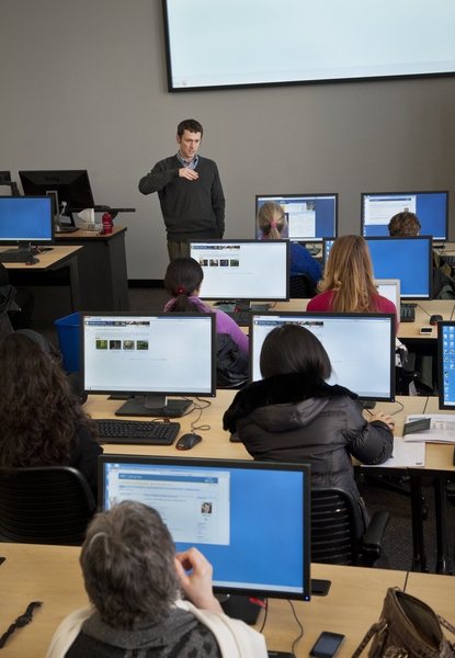 Back view of half a dozen people sitting at long tables, each with a computer monitor in front of them. An instructor is at the front of the class