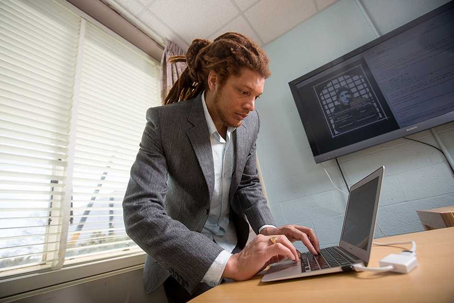 Fox Harrell types at laptop on table. A window is behind him and a tv is mounted on adjacent wall.