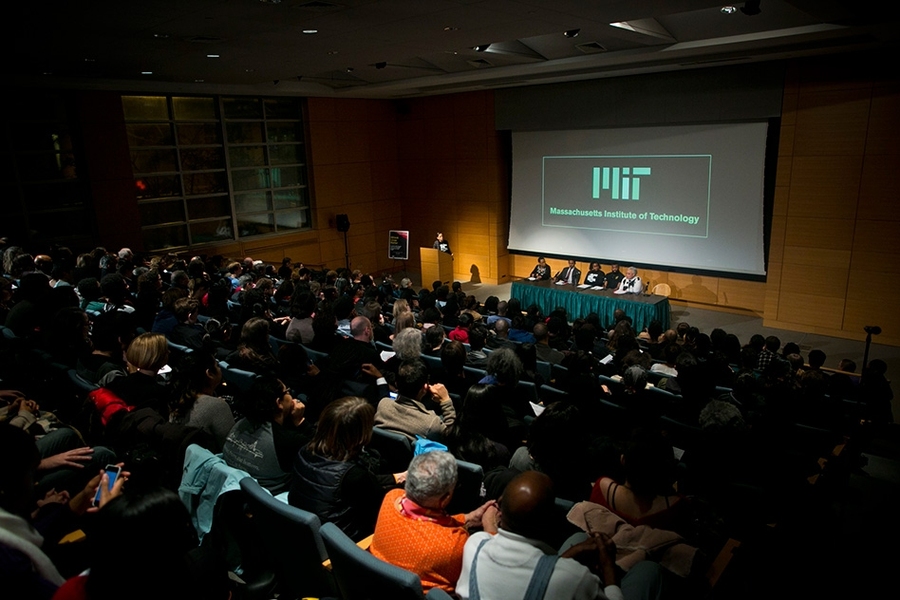 A wide shot image of filled auditorium and five speakers seated at stage
