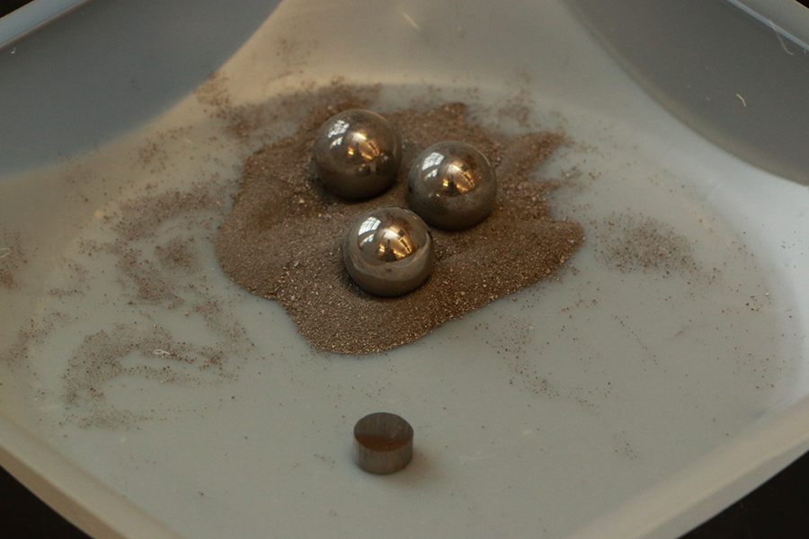 A compacted metal alloy pellet sits next to as-milled tungsten-chromium iron metal powders in a boat used for weighing the metals. The steel balls are used to deform the metals in a high-energy ball mill.