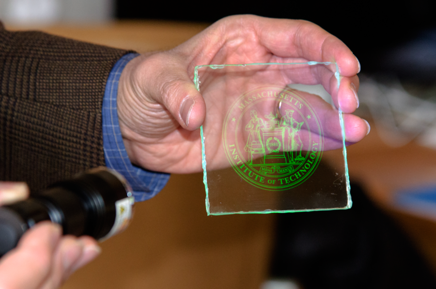 Professor Vladimir Bulović demonstrates how the MIT logo, patterned in thin layers of quantum dots, appears on a seemingly clear piece of glass when an LED light shines on it causing the quantum dots to glow. The thin layers of quantum dots were printed on a 3-inch square piece of glass using inkjet printing.