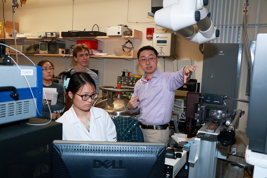 Glass for advanced photonics: Graduate student Lan Li, foreground, left, and Professor Juejun Hu explain summer research on glass for advanced photonics in Hu’s lab. At rear are 2015 MPC-CMSE Summer Scholars Zhenni Lin and Bart Machielse.