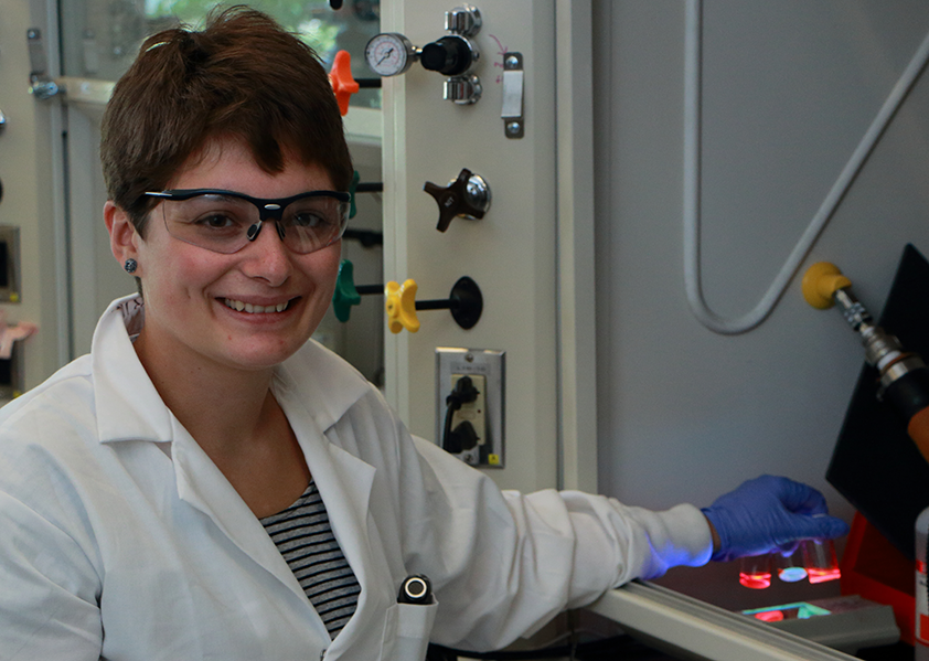 Summer Scholar Katharine Greco holds bottles of core/shell quantum dots over an ultraviolet light to highlight their colors, which glow under UV light. Greco spent the summer synthesizing up to 16 samples of core/shell quantum dots each week in a glovebox in the Tisdale Lab at MIT. Greco used cadmium oleate and octanethiol precursors for her cadmium sulfide shell quantum dot synthesis. 