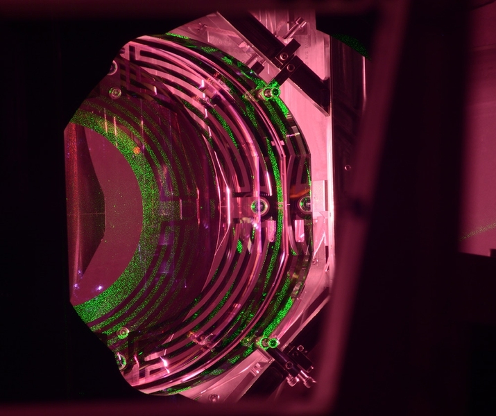 Roughly half of the 100-pound cylindrical Advanced LIGO optic that senses gravitational waves can be seen here. The green light comes from a laser used to help bring the system into an operational state. The maze-like pattern is part of the system to push, twist, and tilt the optic to hold the system in alignment.